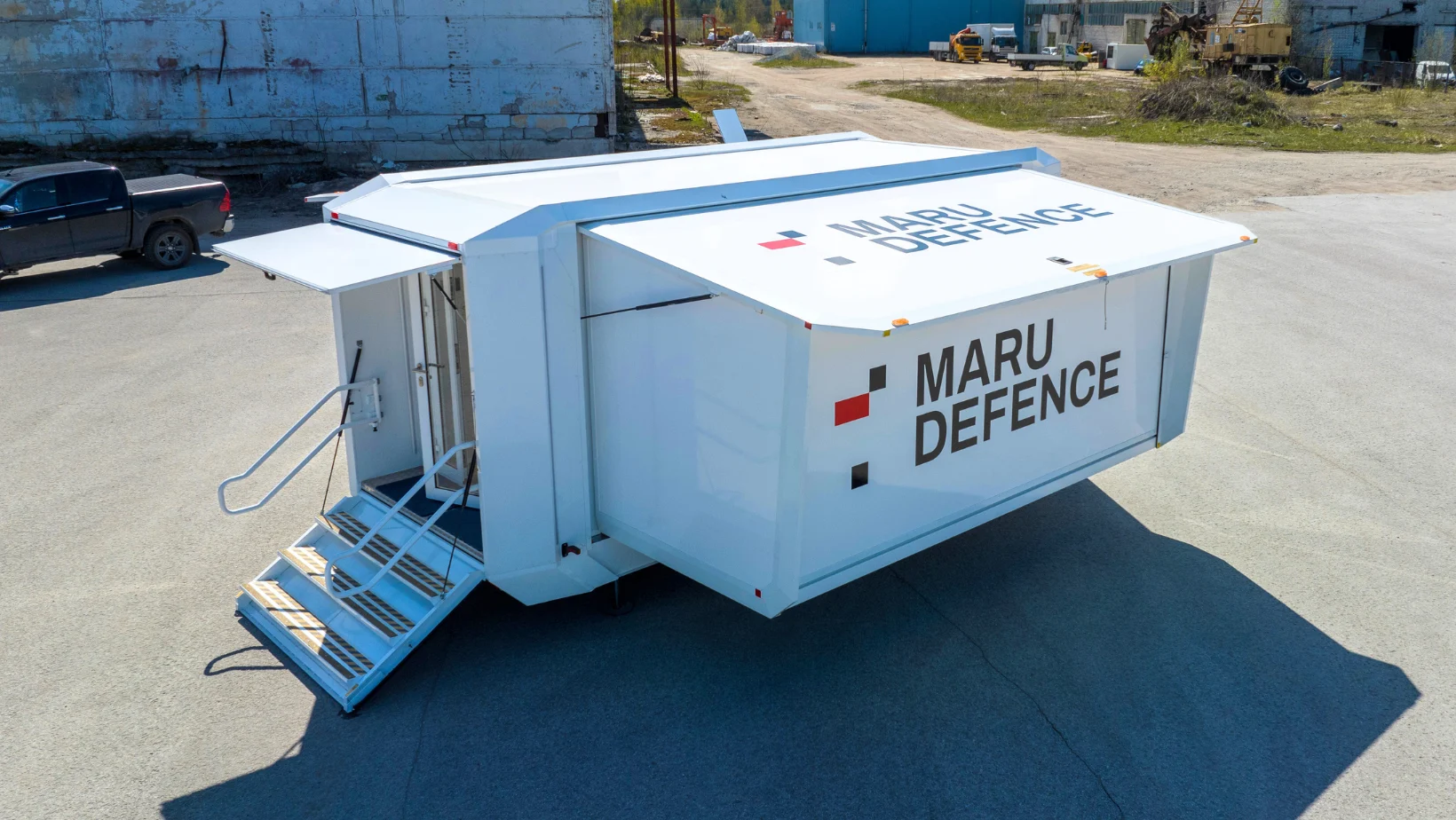 A white Maru Defence mobile trailer with an extended side panel and stairs parked in a lot.