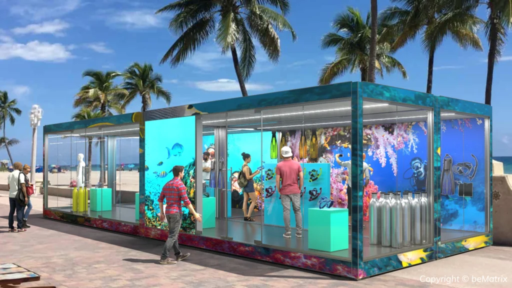 A modular glass pop-up structure decorated with underwater imagery displays scuba diving gear on a beach.