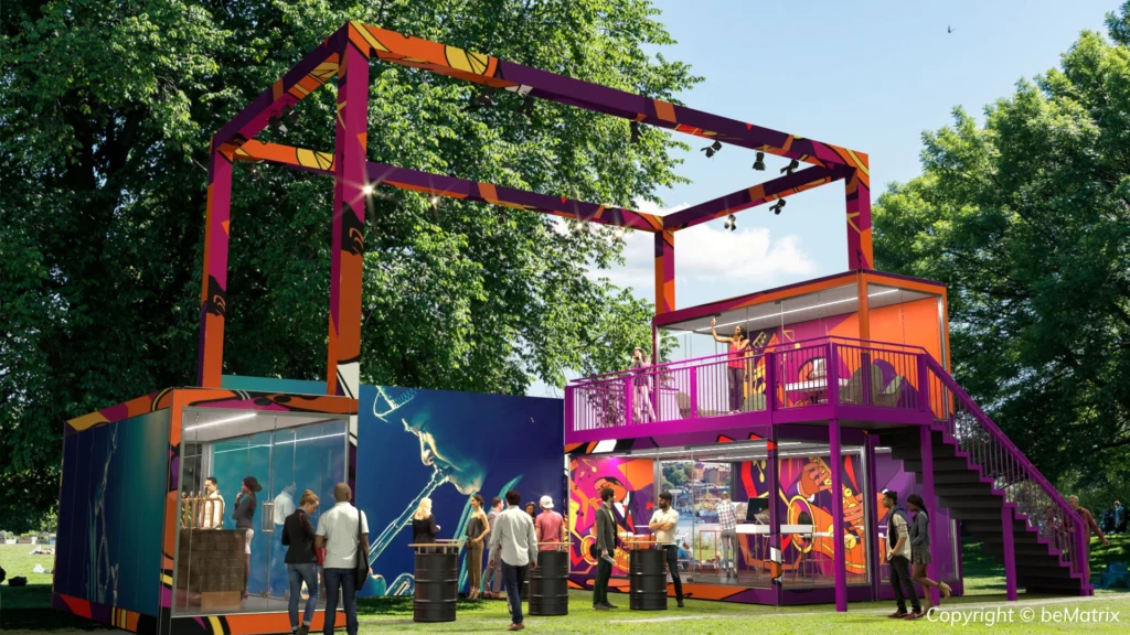 A colorful, two-story modular event structure with an outdoor deck and staircase set in a park.