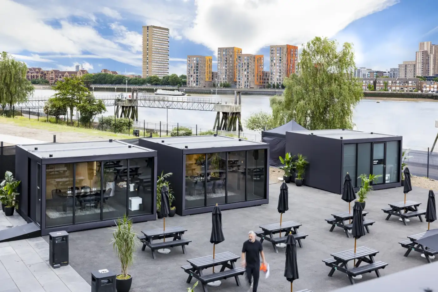 Outdoor patio area with modern glass-walled office pods, picnic tables, and a view of the river and city skyline.