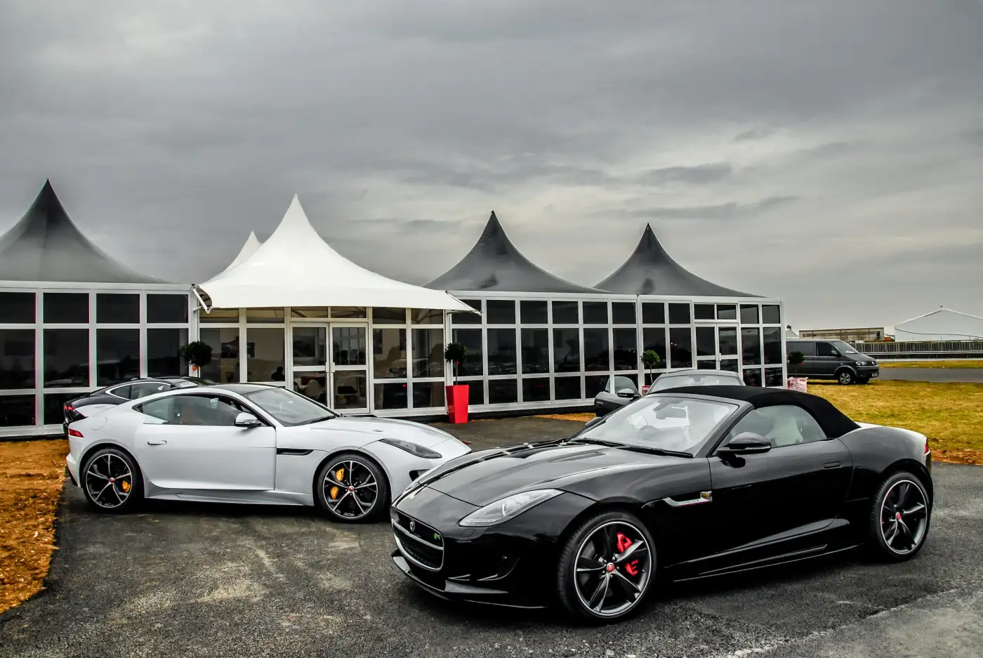 A white and a black Jaguar F-Type parked in front of white event marquees under a cloudy sky.